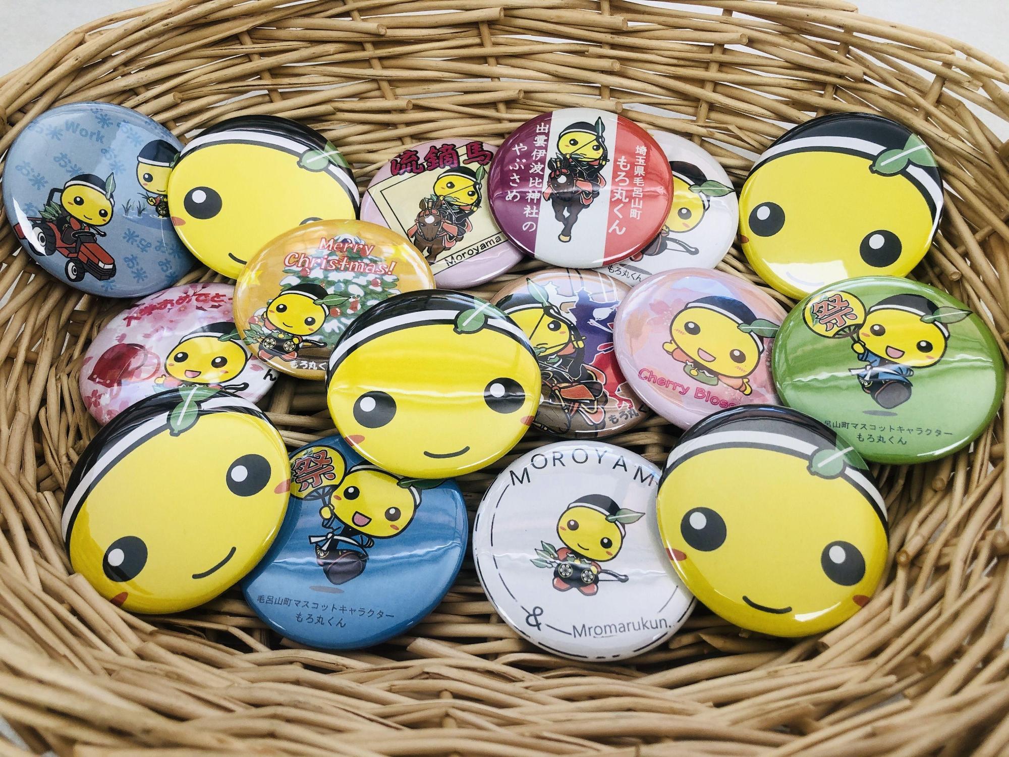 色々なイラストが描かれた毛呂山町キャラクター「もろ丸くん」の缶バッチが竹かごに入っている写真