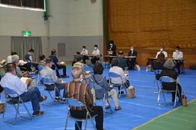 人と人との感覚を開け設置された椅子に座っている参加者とステージ下に置かれた長机の席に2名ずつ座り、黒いスーツ姿の方が立って話をしている様子の写真