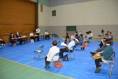 ステージ下に設置された長机の席に参加者の方を向き座っている関係者、うち1名が席の横に立ち、マイクを持って話をしている写真