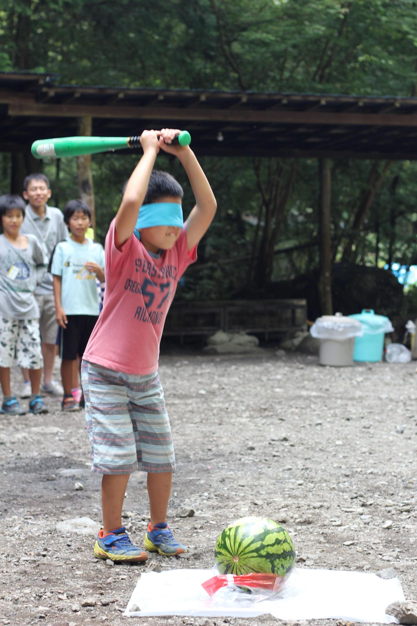 後ろで他の子ども達や大人が見守るなか、赤いシャツを着た男の子が目隠しをして下に置いてあるスイカに向かって野球のバットを振り下ろそうとしている写真