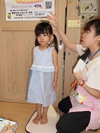 真っ直ぐ前を向いて身長計に乗っている女の子と身長測定をしている保健師さんの写真