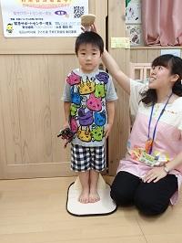 おもちゃを右手に持ったまま、真っ直ぐ前を向いて身長計に乗っている男の子と身長測定をしている保健師さんの写真