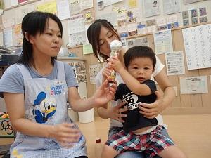 母親に膝抱っこされた男の子が、左に座っているエプロンを着用した女性スタッフが持っていたおもちゃのマイクを取り上げている写真