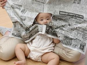 母親に膝抱っこされた赤ちゃんが、母親が両手で持っている新聞紙の破られている隙間から顔を覗かせている写真