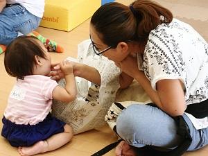 座っている母親が破られている新聞紙の穴から右手を入れて、向かい合って座っている子供の口のあたりを触っている写真