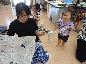 座っている母親が左手で持っている破れた新聞紙の端を、水玉のシャツを着た女の子が笑顔で持っている写真