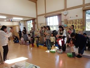 外国人の先生と参加者親子の皆さんが向い合って立っており、両手を合わせる同じ動作をしている「英語で遊ぼう」の様子の写真