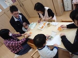 エプロン姿の保育士の横で、クレヨンを使いお絵かきするお母さんや男の子の写真