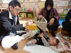仰向けの赤ちゃんがお父さんとお母さんが持つ新聞紙に触ろうと手を伸ばしている写真