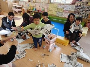 先生の真似をして一人の男の子が新聞を破って楽しんでいて、周りでも他の親子が新聞紙で遊んでいる写真