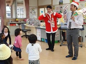 サンタクロースの衣装を着たユーゴ先生とサンタクロースの帽子をかぶったポール先生、向かい合って立っている子供たちの写真