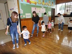オーバーオールを着たお母さんと子供が先頭に手を繋ぎ、室内を歩いている写真