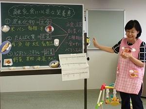 移動式の黒板を使って、離乳食に必要な栄養素を説明しているピンク色のエプロンをつけた栄養士さんの写真