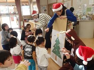 サンタさんのお赤い帽子を被った先生方、トナカイさんに変身した先生などと一緒に、クリスマスツリーの絵が貼ってあるボードに子ども達がシールを貼って飾り付けをしている写真。