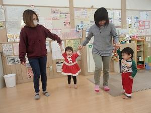 左右大人の女性と手をつないでいる赤いサンタクロースのワンピースを着た女の子と左端の女性と手をつないでいるサンタクロースの赤いズボンを履いている男の子の写真