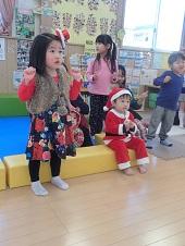 両手をグーと握って腕を曲げたポーズをし、前方を真剣な表情で見ている赤い帽子被った女の子、ブロックに座って前方を見ているサンタクロースの赤い服を着た男の子、その後ろに笑顔で前方を見ている子ども達の写真