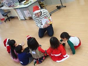 トナカイの帽子を被った外国人の先生が、6人の子ども達に絵本の読み聞かせをしている写真