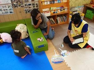 ぬいぐるみが置いてある遊具スペースに女の子が座り、母親と対面で座っている黄色のエプロンを着用した女性スタッフがスタンプを押している写真