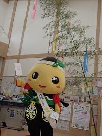 笹飾りの前に立つ〇(丸)のカードを右手に持って揚げているもろ丸くんの写真