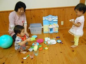ピンク色の女性スタッフが見守るなか、前のよだれかけを掛けた子どもが座っておままごとセットを触っているのを、右側で立って見ている女の子の写真