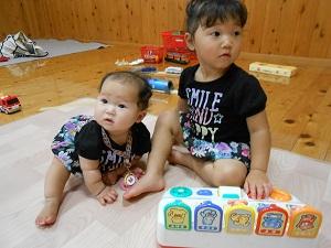 マットの上に座っている右側の大きな女の子が右を見て左手でおもちゃを触り、左側の小さな女の子は左側を見ているお揃いの洋服を着た2名の女の子の写真