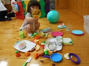 カメラに向けて黄色の透明の容器を向けている女の子がしゃがんだ床に、散らかしたおまままごとセットの写真