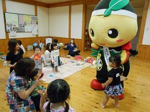 公民館の床に座って小さな男の子を膝に乗せている母親の所に、女の子がゆるキャラ「もろ丸くん」の手を引っ張って連れてきた写真
