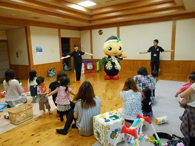 ゆるキャラ「もろ丸くん」を中央に両側に黒色の服を着た2名のスタッフが両手を広げて踊りを披露しているのを、床に座って見ている親子の写真