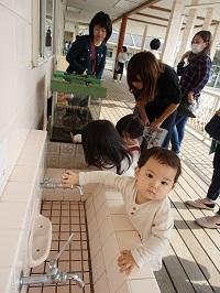 手洗い場で蛇口に手を伸ばしてカメラ目線で見ている小さな子ども、子ども達の様子を見守っているお母さん方の写真