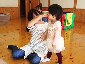 床に座っている女性が左手で女の子の右手を握り、右手をグーにして女の子の顔に近づけて見せている写真