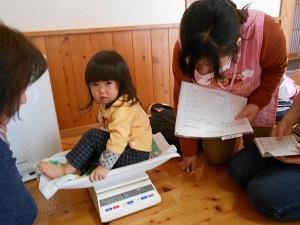 体重計に座ってる女の子、記録用紙を手に持って体重計のメモリを読んでいる保健師の写真