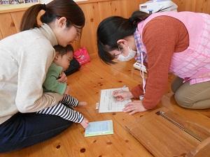 赤ちゃんを膝の上に抱いている母親と記録用紙に書き込みをしている保健師の写真