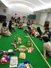 子どもを膝の上に座らせている参加者の皆さんと保育ボランティアさんがおもちゃやぬいぐるみが沢山置かれている緑色のマットの上に座っている写真
