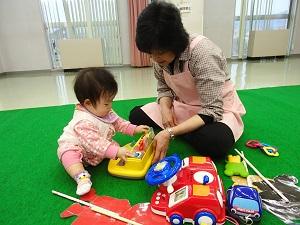 赤ちゃんが黄色いおもちゃで遊んでおり、保育ボランティアさんが赤ちゃんの傍に座って一緒に遊んでいる写真
