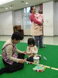 赤ちゃんを抱っこしてあやしている保育ボランティアさん、おもちゃで遊んでいる女の子の傍に座って一緒に遊んでいる保育ボランティアさんが写っている写真