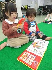 アンパンマンのおもちゃを手に持って遊んでいる女の子と横に座っている小さい男の子。音楽のなる絵本が置かれている写真