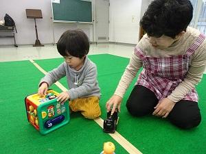 正方形の形で色々な仕掛けのあるおもちゃで遊ぶ男の子。その横で見守っている保育ボランティアさんの写真