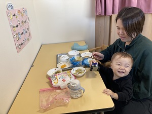 おもちゃを持ち笑顔の幼児と、試食のタイミングを見守る母