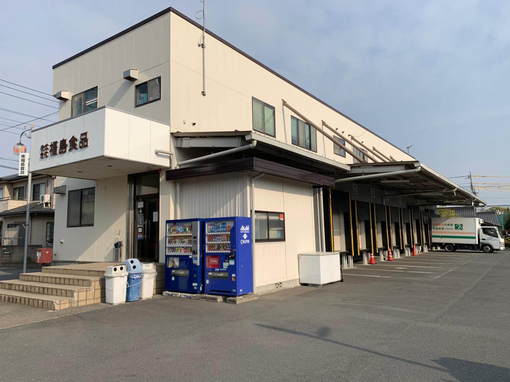 白い壁に2階建ての建物で、奥の倉庫にトラックが停まっている福島食品の写真
