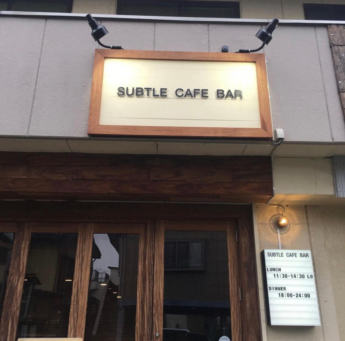 建物に設置された「subtle cafe bar サトルカフェバー」の看板にライトが照らされているサトルカフェバーの外観写真