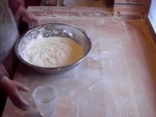 麺打ち台の上に、小麦粉等のうどんの材料を入れた銀色のボールと、右手で水の入った計量カップに手を伸ばしている様子の写真
