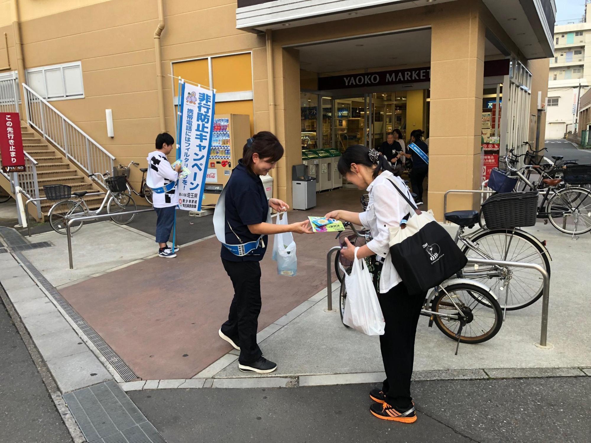 スーパーマーケットの入り口付近で非行防止キャンペーンののぼり旗をもって立っている男性と、キャンペーンのチラシを配っている女性の写真