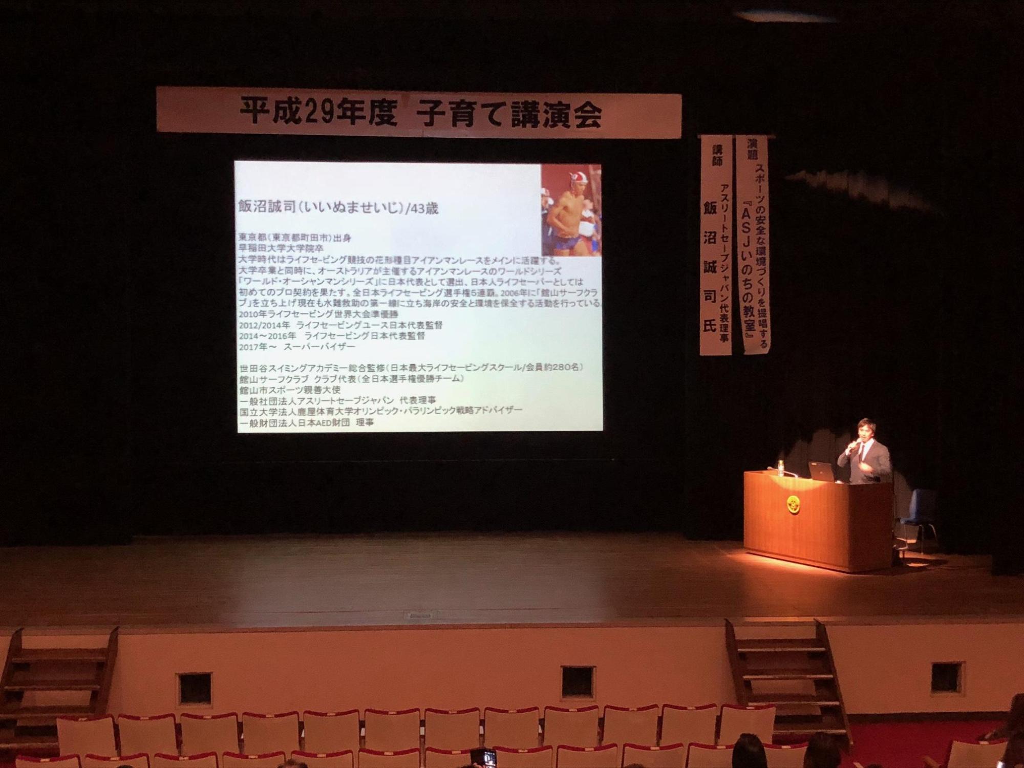 舞台上のスクリーンに映し出された講演会の資料と、舞台の右側で話をしている講師の写真
