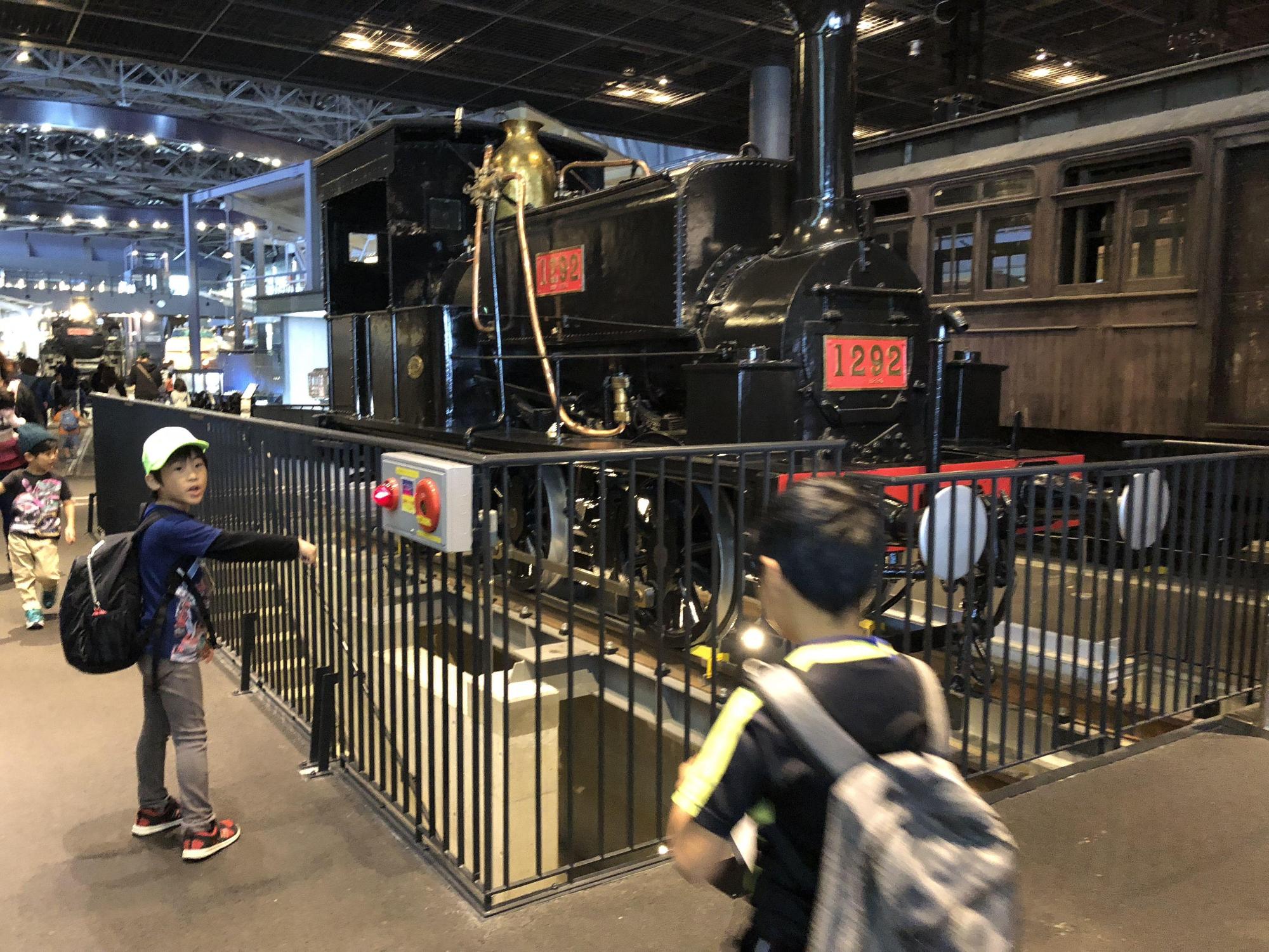 展示されている黒色のボディの蒸気機関車を右側から歩いてくる男の子に向かって指差しながら話しをしている男の子の写真
