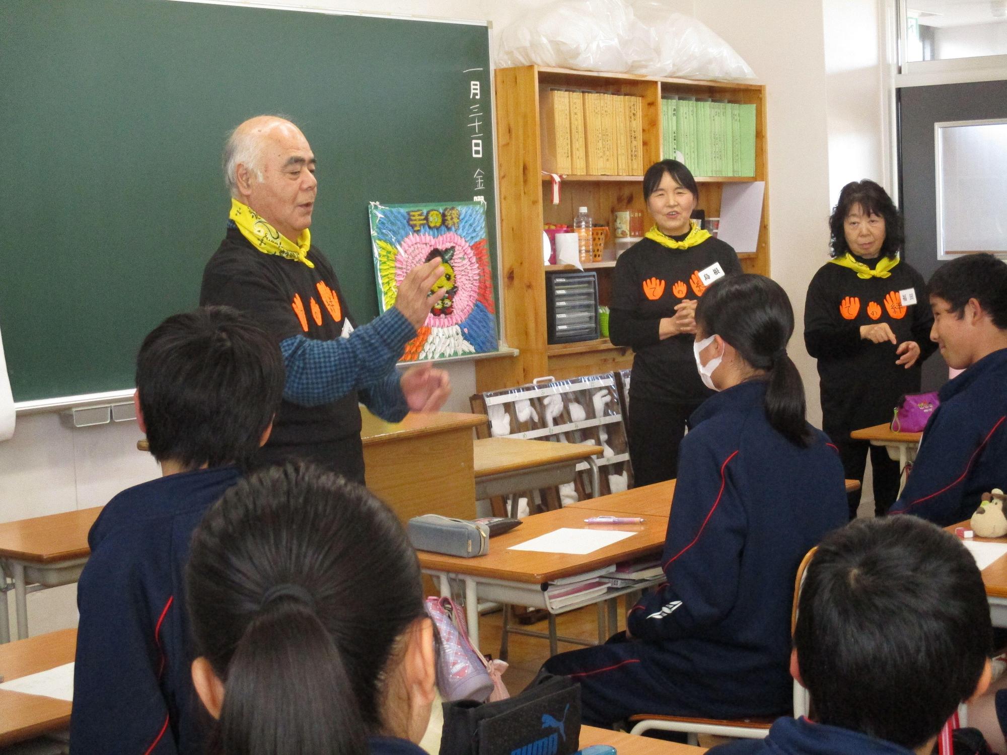 首に黄色いバンダナを巻いた男性が、生徒の前で手話をしている写真