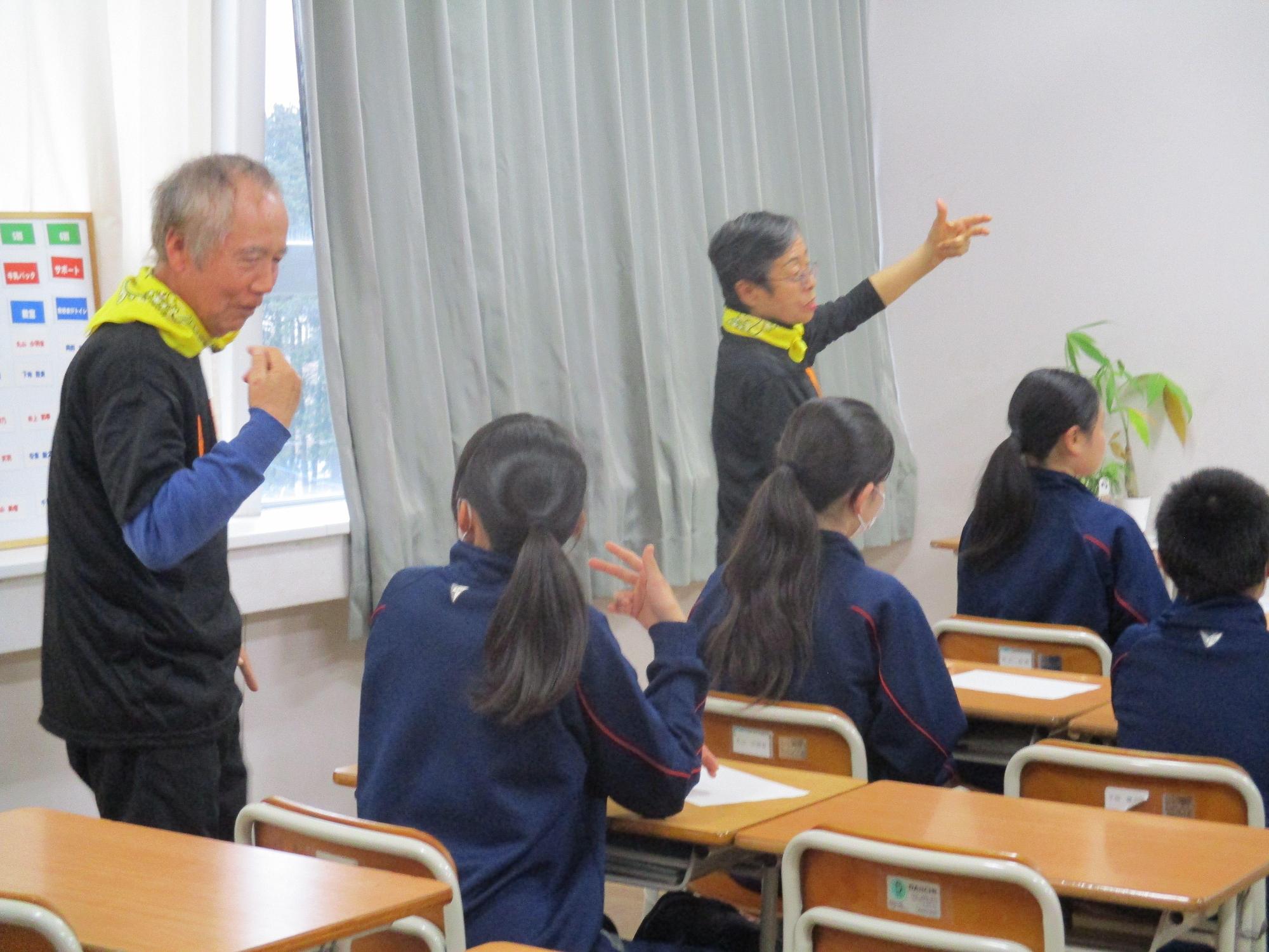 黄色いバンダナを首に巻いた男性と女性が、生徒の横で片手をあげて手話をしている写真
