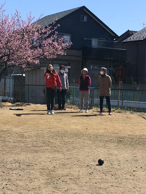第九団地公園内のピンク花が咲く木の前で、赤い服を着た女性が黒いボールを投げ、それを見守る3人の参加者達の写真