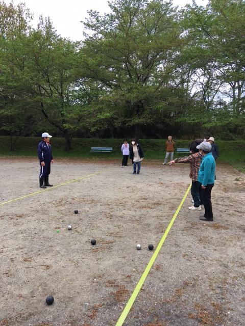 緑の木が生い茂る岡本団地ふれあい広場にて、黒や銀色のボールを使いペタンクを楽しむ参加者達の写真