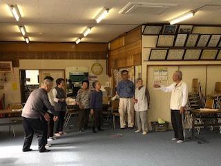 沢田公会堂内で、ペタンクの説明をするクリーム色のベストを着た男性と熱心に話を聞く参加者達の写真