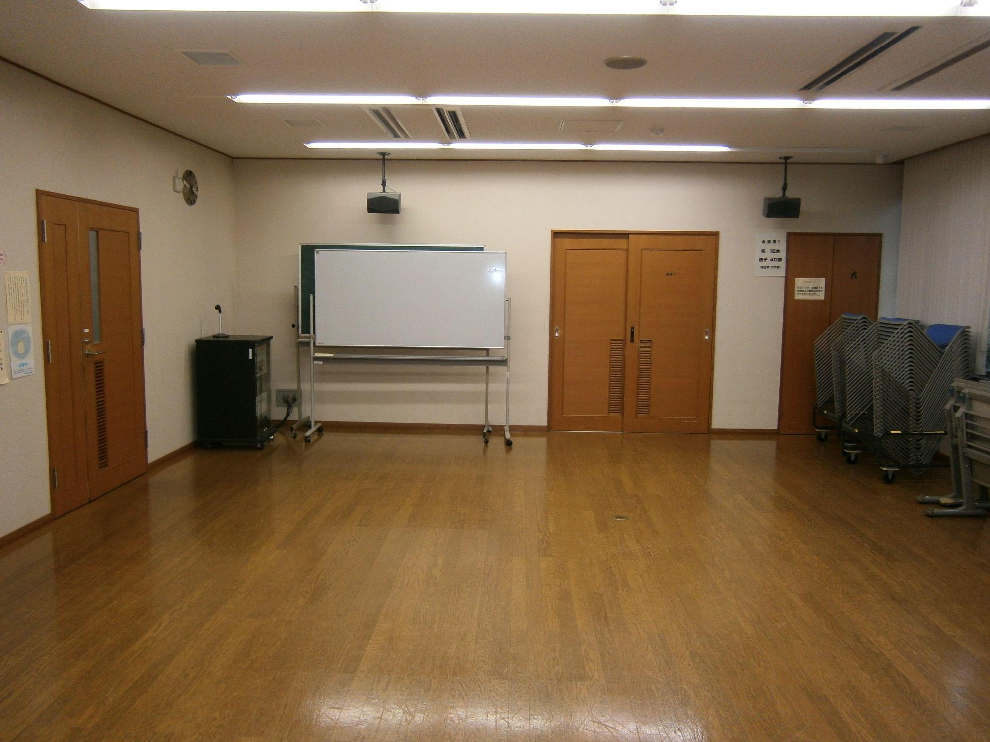 左側に出入口のドアがあり、室内の奥に可動式の黒板とホワイトボードが置かれ、右壁側に椅子と長机が積み上げられている会議室の写真
